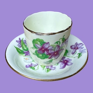 Peut inclure: Un ensemble tasse et soucoupe vintage. La tasse et la soucoupe sont blanches avec une bordure dorée. Le motif représente des violettes violettes avec des feuilles vertes. La tasse est cylindrique et repose sur une soucoupe assortie.