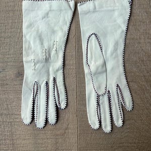 May include: A pair of off-white gloves with black and red stitching along the edges and seams. The gloves are made of a soft material and have a classic design. The gloves are laid out on a wooden surface.