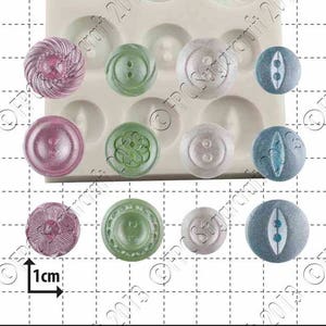 May include: A silicone mould with 14 different button shapes. The mould is white and has a 1 cm ruler for scale. The buttons are in various colours and designs, including swirls, flowers, and two-hole buttons.