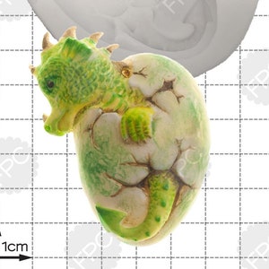 May include: A silicone mold for baking a green dragon emerging from a cracked egg. The mold is white and shows the dragon's head, wings, and tail. The dragon is green with yellow accents and is shown in a cracked egg.