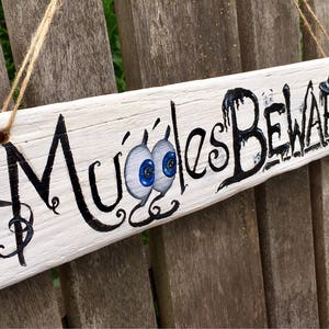 May include: A rectangular, white wooden sign with the words "Muggles Beware" in black, whimsical lettering. The sign features cartoon eyeballs and a witch's hat. It hangs by a twine string, perfect for Halloween or a spooky theme.