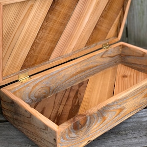 May include: Open wooden box with a hinged lid. The box is constructed from light and dark brown wood planks arranged in a diagonal pattern. The interior is visible, and the box is empty. The hinges are brass-colored.