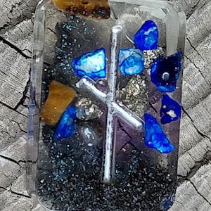 May include: A rectangular resin pendant with a silver metal rune symbol. The pendant is filled with blue and brown stones, silver flecks, and black glitter. The background is a weathered wood surface.