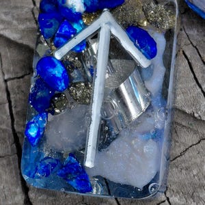 May include: A rectangular resin pendant with a white rune symbol, blue stones, silver metal, and gold flecks. The pendant is filled with a clear resin, creating a layered effect. The blue stones are a vibrant contrast to the other elements.