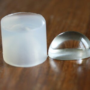 May include: A clear, round, glass dome sits next to a clear, cylindrical, plastic mould. Both objects are on a wooden surface.
