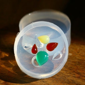 Könnte beinhalten: Eine transparente Silikonform mit fünf tropfenförmigen Vertiefungen. Vier Vertiefungen sind mit farbigem Harz gefüllt: rot, grün, gelb und dunkelrot. Die Form steht auf einer Holzoberfläche.