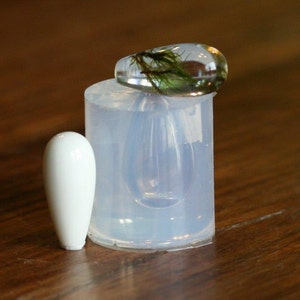 Puede incluir: Un molde de silicona transparente para hacer joyas de resina. El molde tiene forma de l&aacute;grima y tiene un peque&ntilde;o objeto cil&iacute;ndrico blanco a su lado. Un colgante de resina transparente con musgo verde en su interior est&aacute; sentado encima del molde.