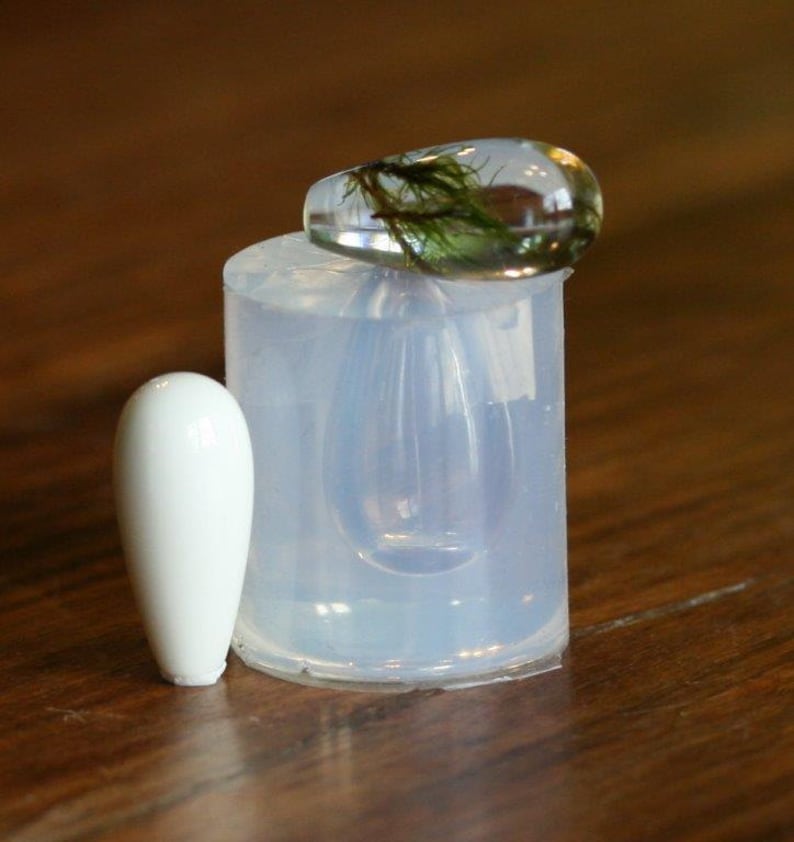 Puede incluir: Un molde de silicona transparente para hacer joyas de resina. El molde tiene forma de l&aacute;grima y tiene un peque&ntilde;o objeto cil&iacute;ndrico blanco a su lado. Un colgante de resina transparente con musgo verde en su interior est&aacute; sentado encima del molde.