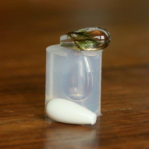 Puede incluir: Un colgante de resina transparente con una peque&ntilde;a planta verde en su interior, sentado sobre un molde de silicona transparente. Tambi&eacute;n se muestra un colgante de cer&aacute;mica blanca.