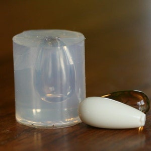 Puede incluir: Un molde de silicona transparente para hacer cuentas de vidrio en forma de l&aacute;grima. Se muestran dos cuentas de vidrio en forma de l&aacute;grima junto al molde, una es blanca y la otra es &aacute;mbar transl&uacute;cido.