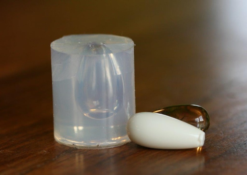 Puede incluir: Un molde de silicona transparente para hacer cuentas de vidrio en forma de l&aacute;grima. Se muestran dos cuentas de vidrio en forma de l&aacute;grima junto al molde, una es blanca y la otra es &aacute;mbar transl&uacute;cido.
