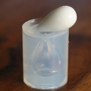 May include: A clear silicone mold for making a teardrop-shaped pendant. The mold is filled with a clear resin and a white teardrop-shaped object is placed on top of the mold.