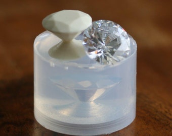 May include: A clear silicone mold for making a diamond-shaped pendant. The mold is filled with a clear resin and a small white diamond-shaped object is sitting on top of the mold.