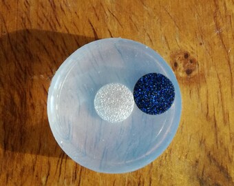 Könnte beinhalten: Eine runde, transparente Silikonform mit zwei kleinen Kreisen im Inneren. Ein Kreis ist mit silbernem Glitzer und der andere mit blauem Glitzer.
