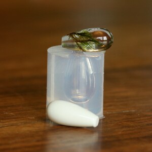 Puede incluir: Un molde de silicona transparente para hacer colgantes de resina en forma de l&aacute;grima. Un colgante terminado con material vegetal verde en su interior est&aacute; sentado encima del molde.