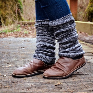 May include: A pair of dark blue denim jeans worn with charcoal gray and white marled knit leg warmers that feature a ribbed texture. The leg warmers are paired with brown leather slip-on shoes that have a rounded toe and a flat black sole.