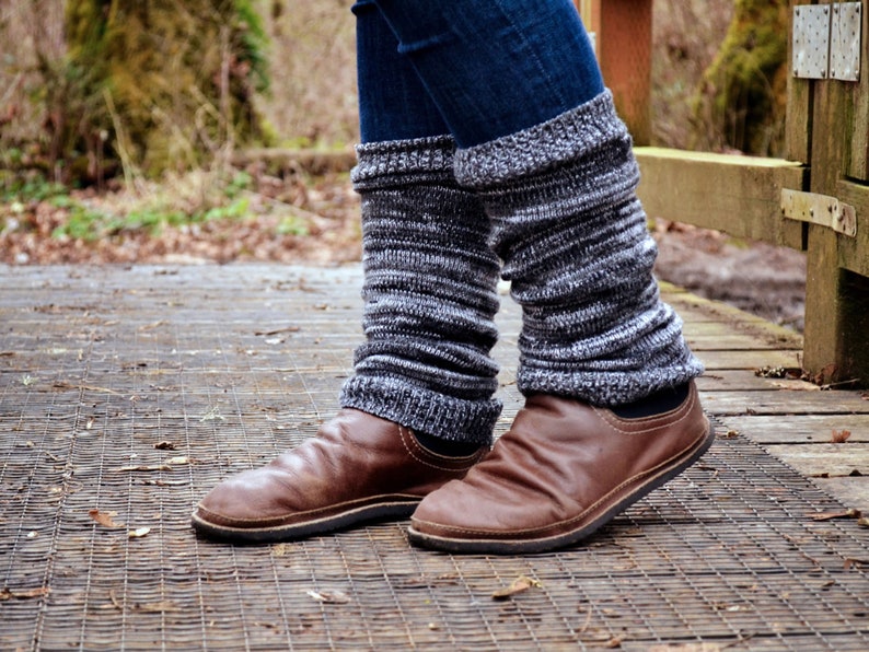 May include: A pair of dark blue denim jeans worn with charcoal gray and white marled knit leg warmers that feature a ribbed texture. The leg warmers are paired with brown leather slip-on shoes that have a rounded toe and a flat black sole.