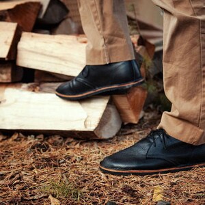 Puede incluir: Un par de zapatos de cuero negro con suela marr&oacute;n. Los zapatos son usados por una persona de pie sobre una pila de madera.