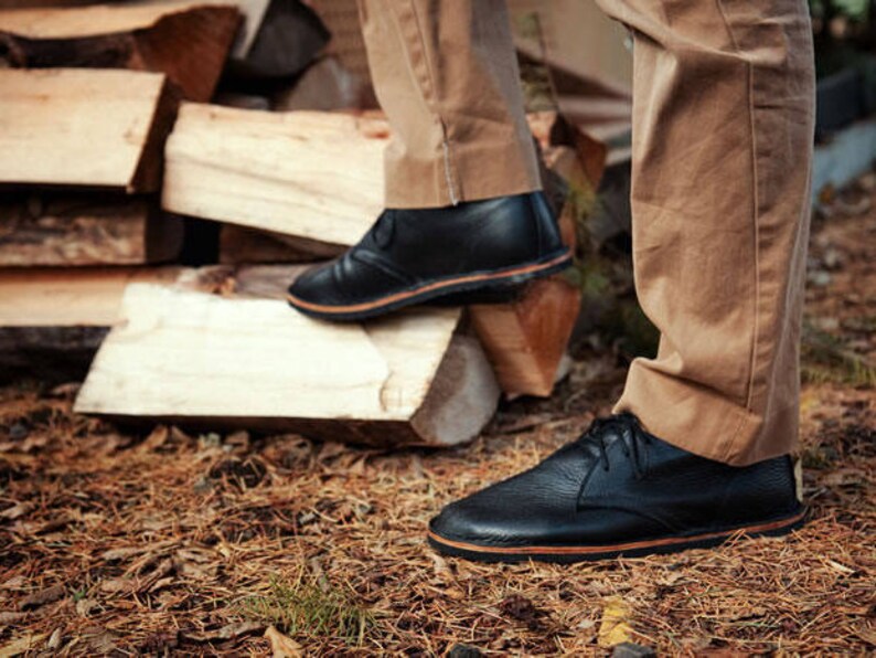 Puede incluir: Un par de zapatos de cuero negro con suela marr&oacute;n. Los zapatos son usados por una persona de pie sobre una pila de madera.