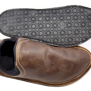 May include: A brown leather slip-on shoe with a black interior lining and white contrast stitching. The shoe is positioned below its black rubber outsole, which features a textured circular pattern and the Vibram brand name embossed on it.