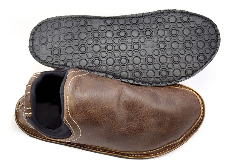 May include: A brown leather slip-on shoe with a black interior lining and white contrast stitching. The shoe is positioned below its black rubber outsole, which features a textured circular pattern and the Vibram brand name embossed on it.