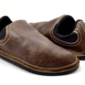 May include: A pair of brown slip-on shoes featuring a textured leather-like upper, contrasting white topstitching, and a black elastic collar. The shoes have a low-profile design with a black rubber sole, providing a simple and functional look.