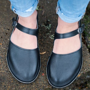 May include: A pair of black leather shoes with a buckle strap and a white trim around the sole. The shoes are being worn by someone wearing blue jeans.