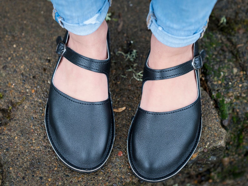 May include: A pair of black leather shoes with a buckle strap and a white trim around the sole. The shoes are being worn by someone wearing blue jeans.