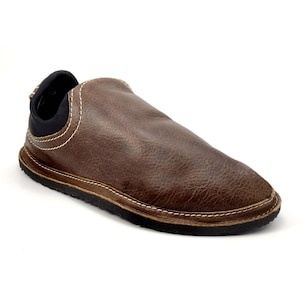 May include: A brown slip-on shoe featuring a textured leather-like upper with visible white contrast stitching along the seams. The shoe includes a black fabric collar around the ankle opening and a black rubber sole with a textured tread pattern.