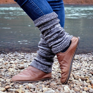May include: A pair of dark blue denim jeans worn with charcoal gray and white marled knit leg warmers that feature a ribbed texture. The leg warmers are paired with brown leather slip-on shoes that have a rounded toe and visible stitching.