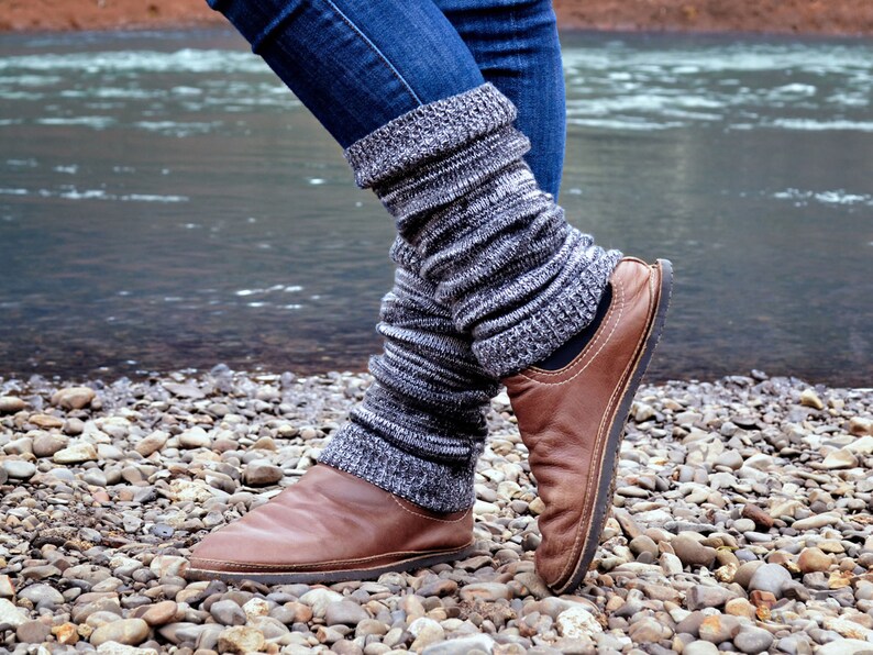 May include: A pair of dark blue denim jeans worn with charcoal gray and white marled knit leg warmers that feature a ribbed texture. The leg warmers are paired with brown leather slip-on shoes that have a rounded toe and visible stitching.