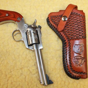 May include: A silver-toned revolver with a wood-grain grip and a matching brown leather holster. The holster features a basket-weave pattern, black braided edges, and two embossed animal head designs, secured by a single silver metal snap.