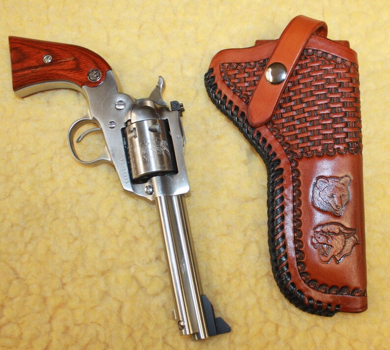 May include: A silver-toned revolver with a wood-grain grip and a matching brown leather holster. The holster features a basket-weave pattern, black braided edges, and two embossed animal head designs, secured by a single silver metal snap.