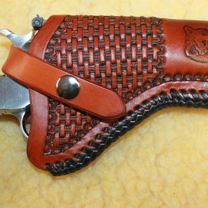 May include: A silver revolver with reddish-brown wood grain grips featuring a circular emblem, housed in a tan leather holster. The holster has a basket-weave pattern, black braided edges, a metal snap closure, and two embossed animal head designs.