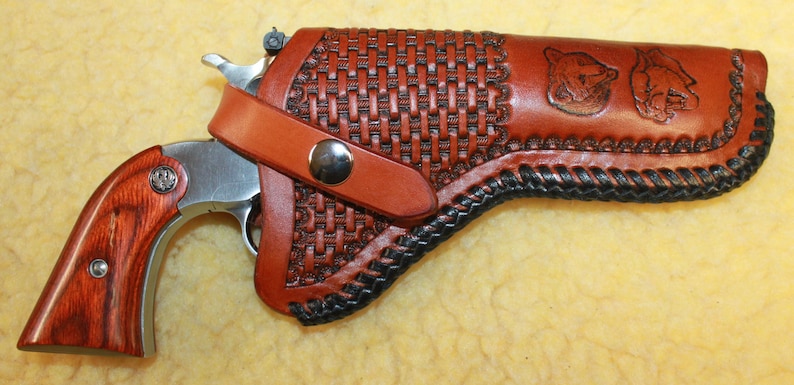 May include: A silver revolver with reddish-brown wood grain grips featuring a circular emblem, housed in a tan leather holster. The holster has a basket-weave pattern, black braided edges, a metal snap closure, and two embossed animal head designs.
