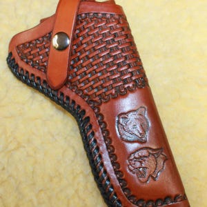May include: A brown leather holster featuring a basketweave pattern, a silver-tone metal snap closure strap, and black lacing along the edges. The surface is embossed with two animal head designs, one resembling a bear and the other a mountain lion.