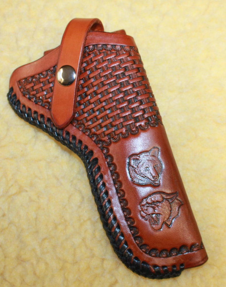 May include: A brown leather holster featuring a basketweave pattern, a silver-tone metal snap closure strap, and black lacing along the edges. The surface is embossed with two animal head designs, one resembling a bear and the other a mountain lion.