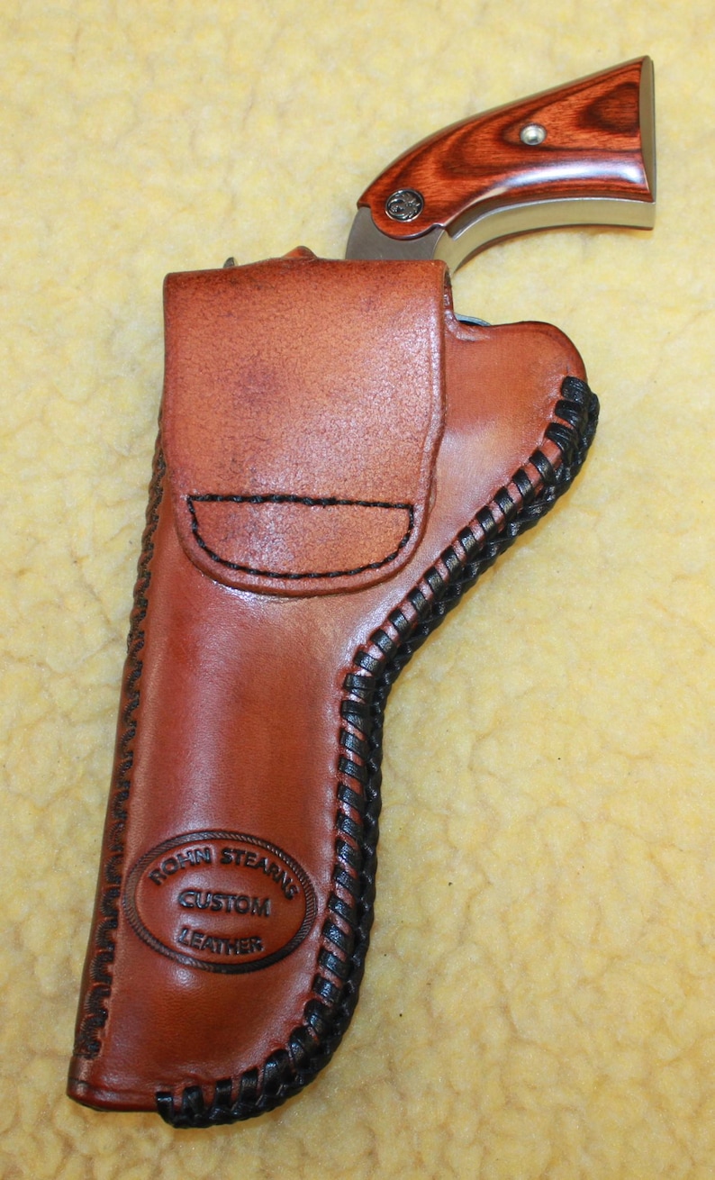 May include: A brown leather holster with black lacing along the edges, featuring a stamped logo that reads Rohn Stearns Custom Leather. A small revolver with a polished metal frame and wood grain grip is partially inserted into the holster.