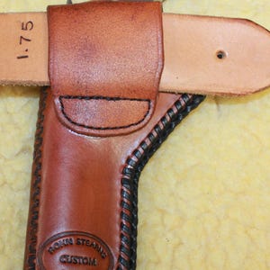 May include: A brown leather holster with black stitching and a stamped logo reading Rohn Stearns Custom Leather. A small revolver with a wood-grain grip is inserted into the holster, which is attached to a light tan belt marked with 1.75 inches.