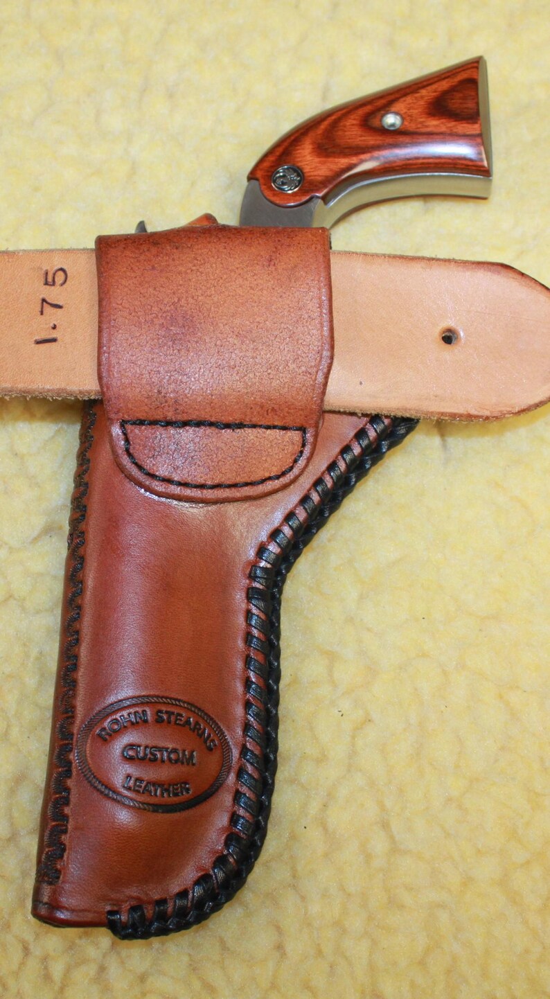 May include: A brown leather holster with black stitching and a stamped logo reading Rohn Stearns Custom Leather. A small revolver with a wood-grain grip is inserted into the holster, which is attached to a light tan belt marked with 1.75 inches.