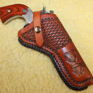 May include: A silver revolver with a wood grain grip is secured inside a tan leather holster. The holster features a basket weave pattern, black braided edge stitching, a metal snap closure strap, and two embossed animal head designs.