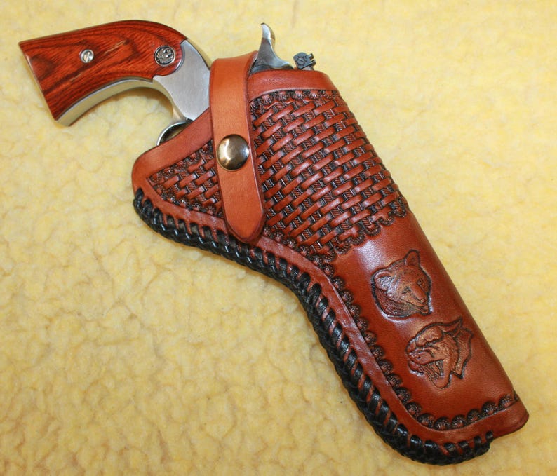 May include: A silver revolver with a wood grain grip is secured inside a tan leather holster. The holster features a basket weave pattern, black braided edge stitching, a metal snap closure strap, and two embossed animal head designs.