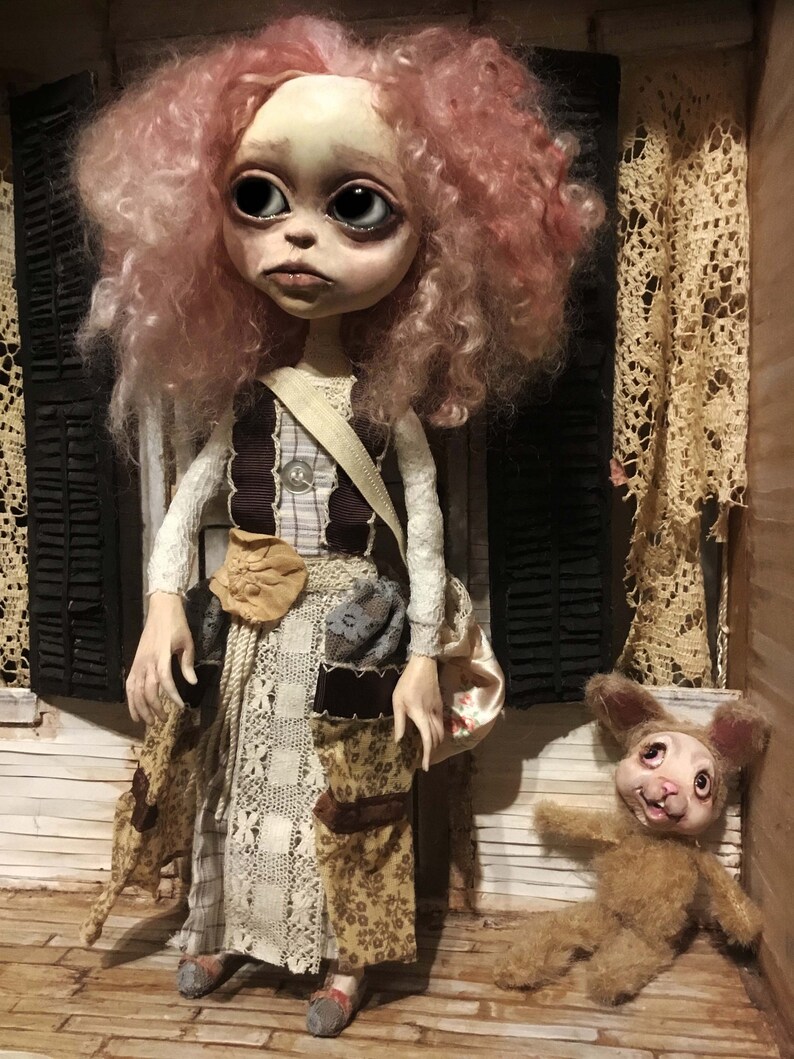 May include: A handcrafted doll with a pale face, large eyes, and pink curly hair. The doll wears a patchwork dress with a beige shoulder bag. A small stuffed bunny lies on the floor nearby. The setting appears to be a miniature room.