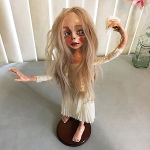 May include: A porcelain doll with long blonde hair wearing a white dress and holding a white tissue. The doll is standing on a wooden base.