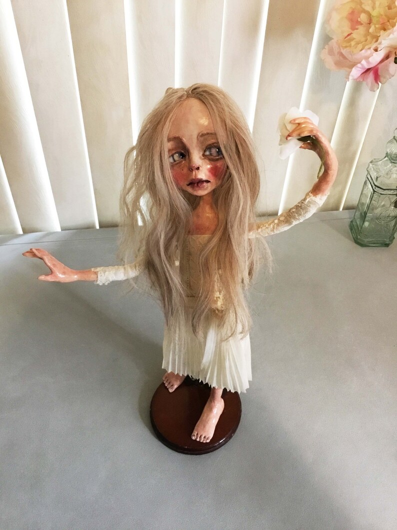May include: A porcelain doll with long blonde hair wearing a white dress and holding a white tissue. The doll is standing on a wooden base.