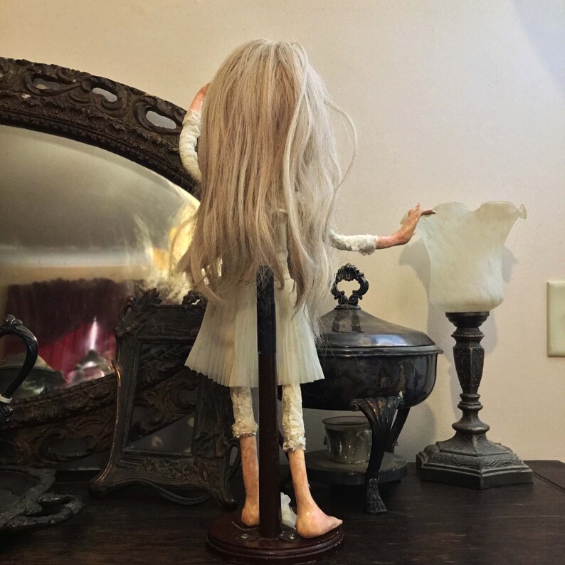 May include: A white porcelain doll with long blonde hair, wearing a white dress and standing on a wooden base. The doll is posed with one arm raised and the other extended to the side. The doll is in front of a mirror and a lamp.