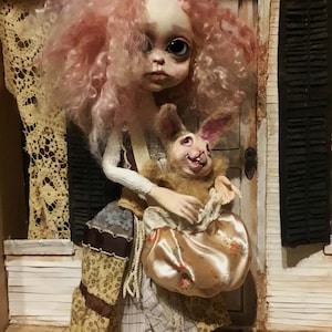 May include: A handmade doll with a pale face, large eyes, and pink curly hair. The doll wears a patchwork dress and holds a plush rabbit with a smiling face and a satin bag with a floral pattern. The doll is posed in a miniature setting.