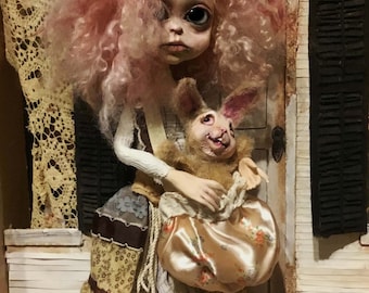 May include: A handmade doll with a pale face, large eyes, and pink curly hair. The doll wears a patchwork dress and holds a plush rabbit with a smiling face and a satin bag with a floral pattern. The doll is posed in a miniature setting.
