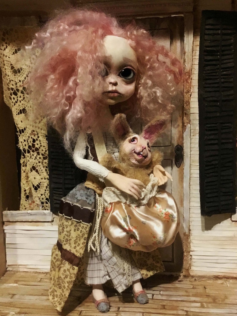 May include: A handmade doll with a pale face, large eyes, and pink curly hair. The doll wears a patchwork dress and holds a plush rabbit with a smiling face and a satin bag with a floral pattern. The doll is posed in a miniature setting.