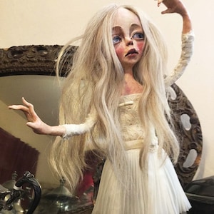May include: A porcelain doll with long white hair, wearing a white dress with a lace bodice and a pleated skirt. The doll has a painted face with blue eyes and rosy cheeks.